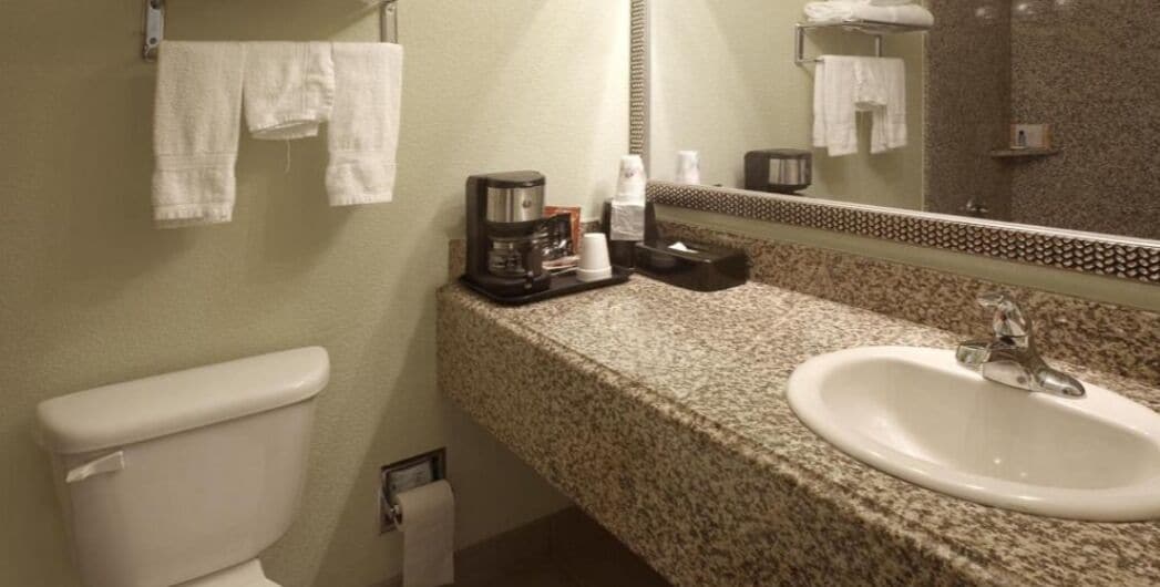 A bathroom with a toilet, sink, and mirror. There are towels hanging on the wall and a coffee maker on the counter.