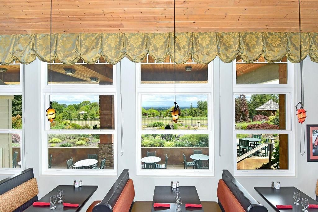 A dining area with multiple windows offering views of a garden and a building. Tables and benches are arranged for seating.