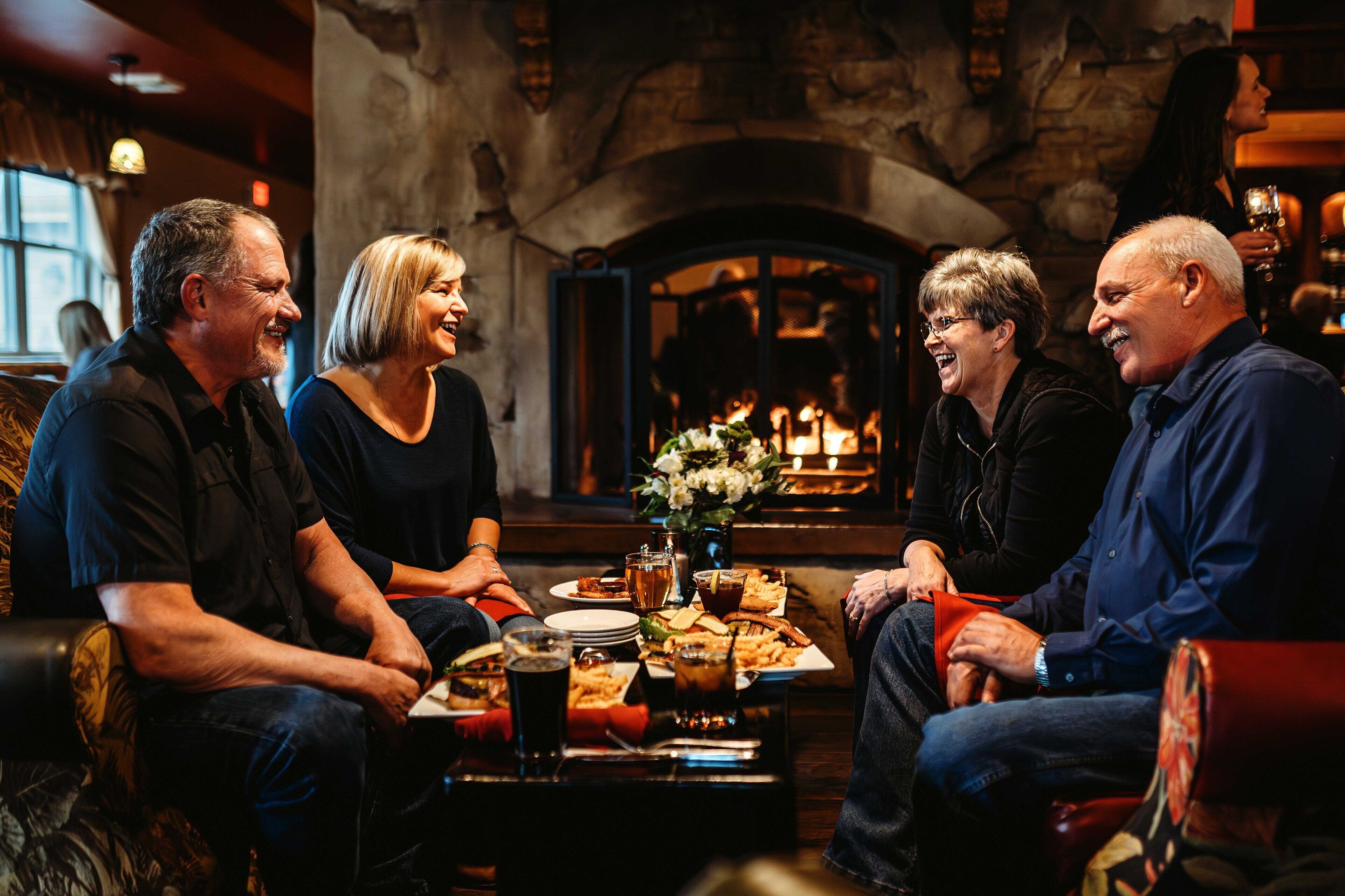 A group of people seated around a table with food and drinks in a cozy room with a fireplace.