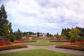 Un jardín ajardinado con un césped central, parterres de flores y un edificio al fondo.