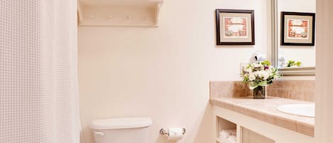 A bathroom with a toilet, a sink with a mirror, a shower curtain, and a shelf with towels.