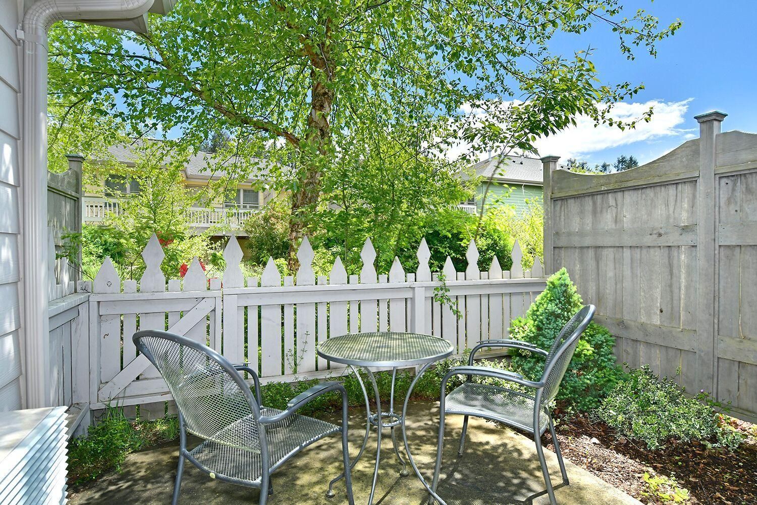 A patio with a metal table and chairs, a white picket fence, and a tree providing shade.