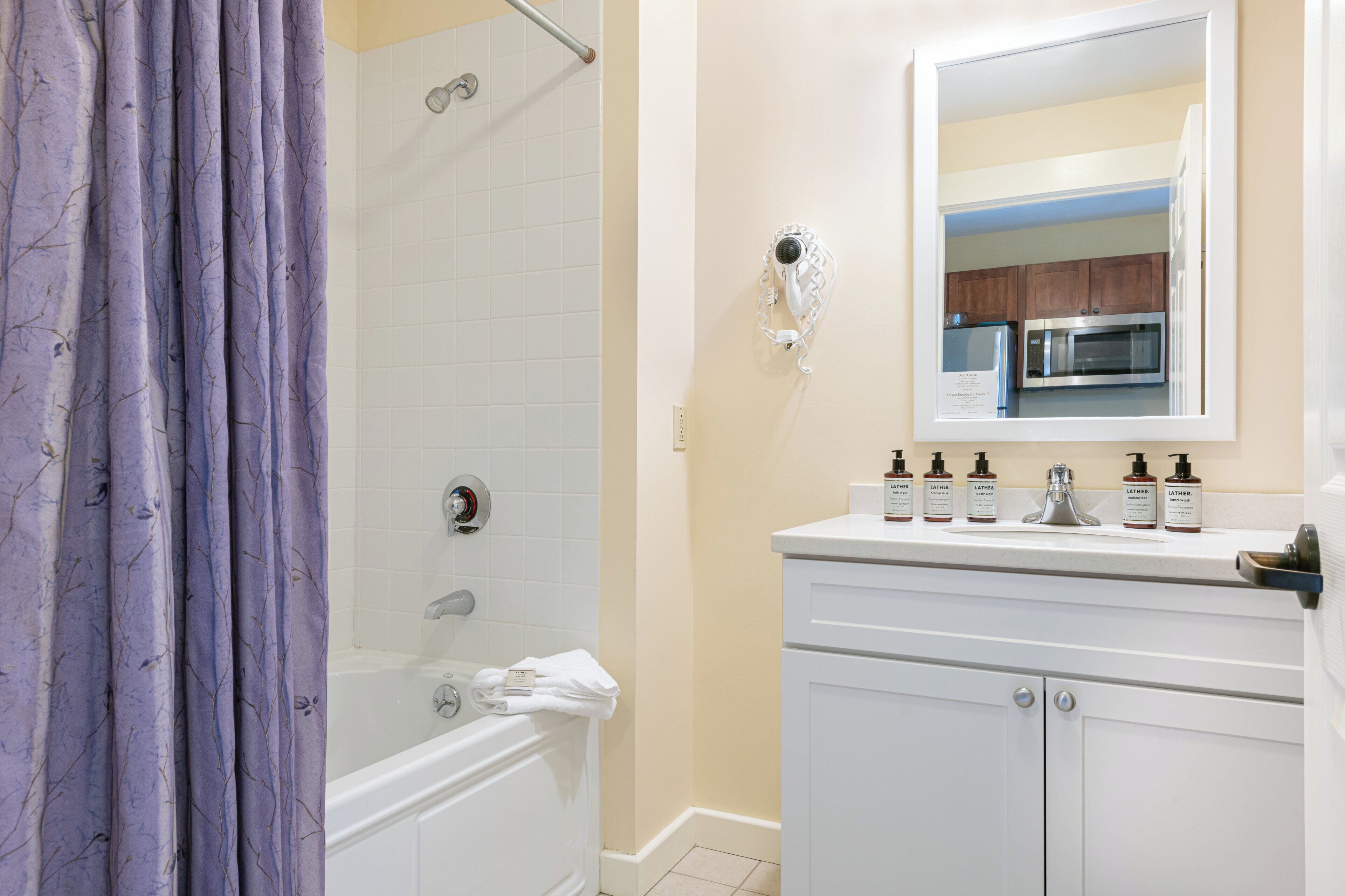 A bathroom with a shower, a sink with toiletries, and a mirror reflecting a kitchen area.