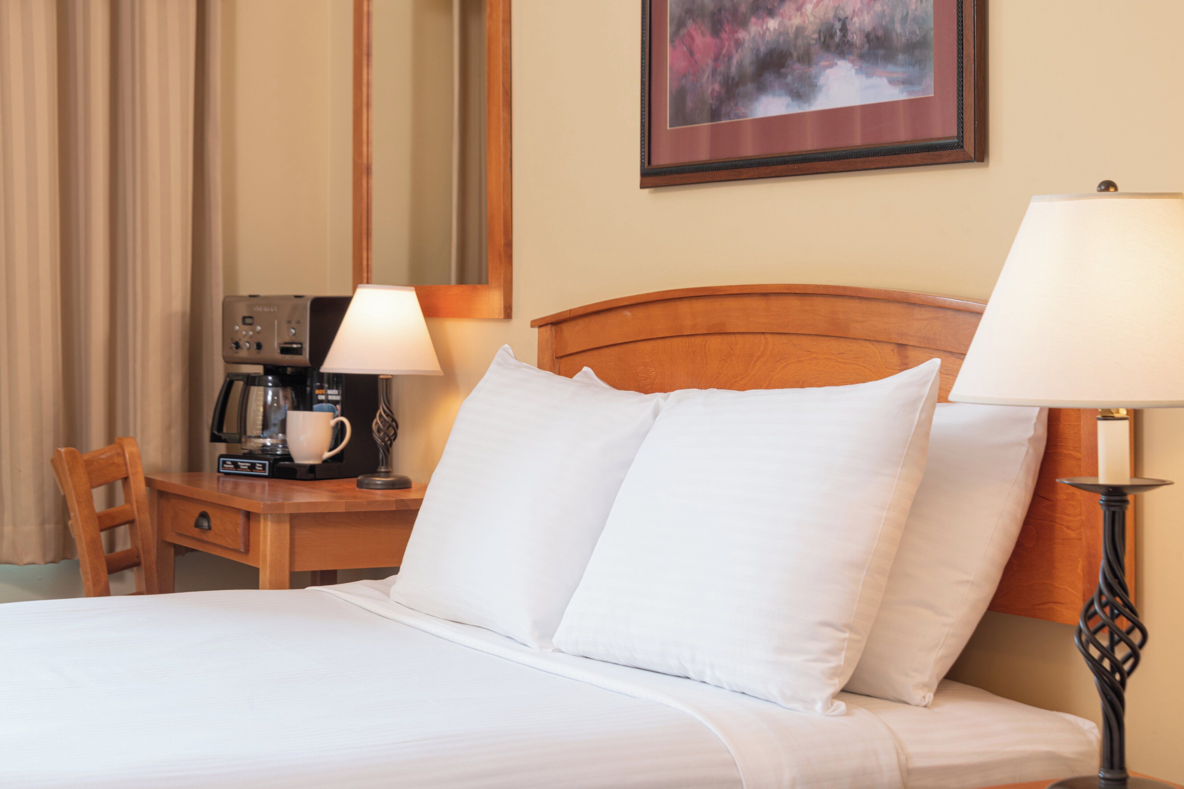 A hotel room with a bed, a desk with a coffee maker, a lamp, and a framed picture on the wall.