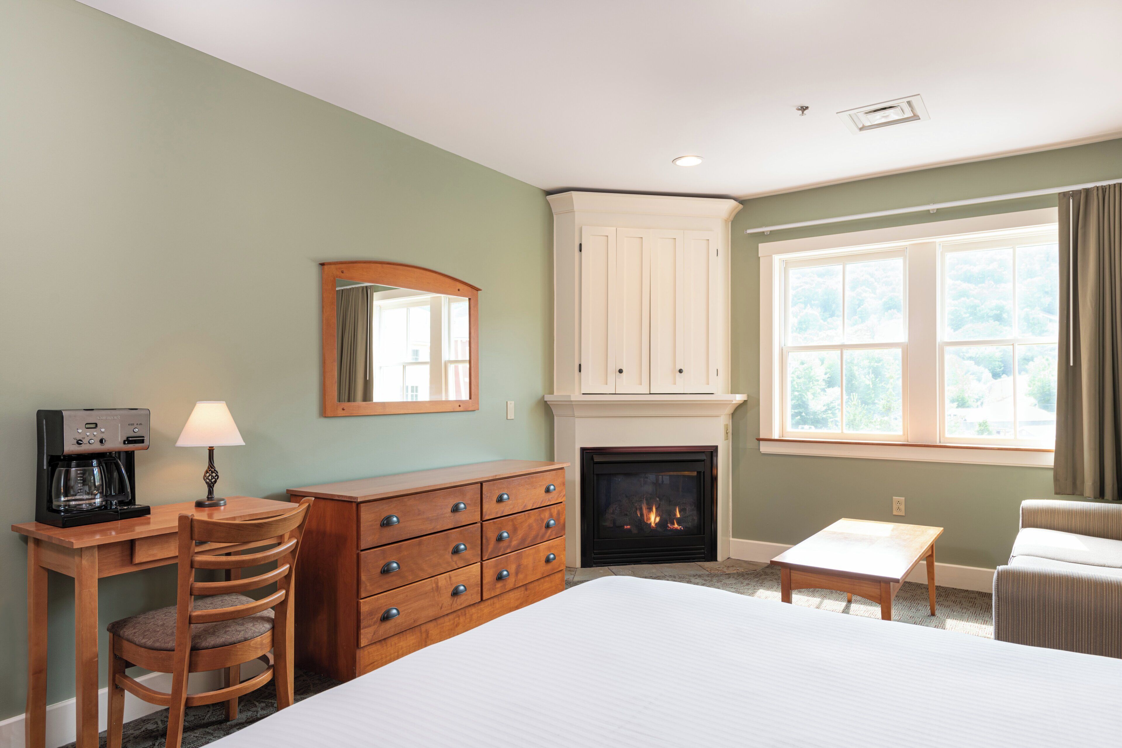 A hotel room with a bed, a desk with a coffee maker, a dresser, a fireplace, and a window with curtains.
