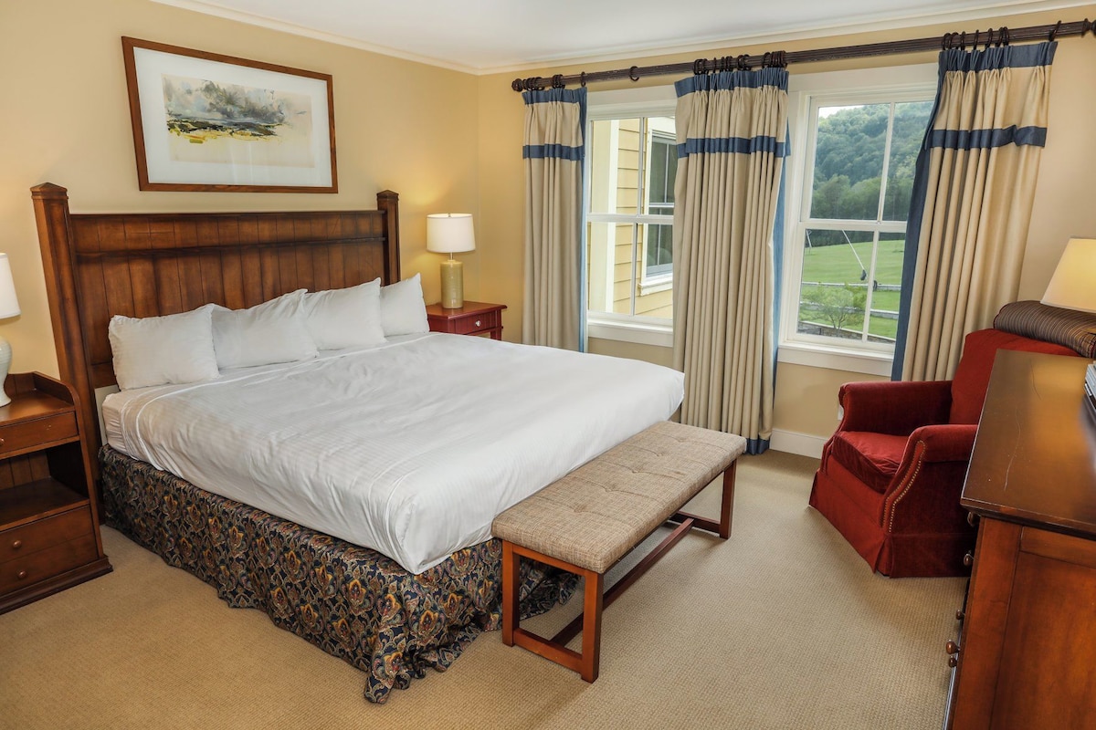 A hotel room with a large bed, a bench, a red armchair, a wooden nightstand, a lamp, and a window with curtains.