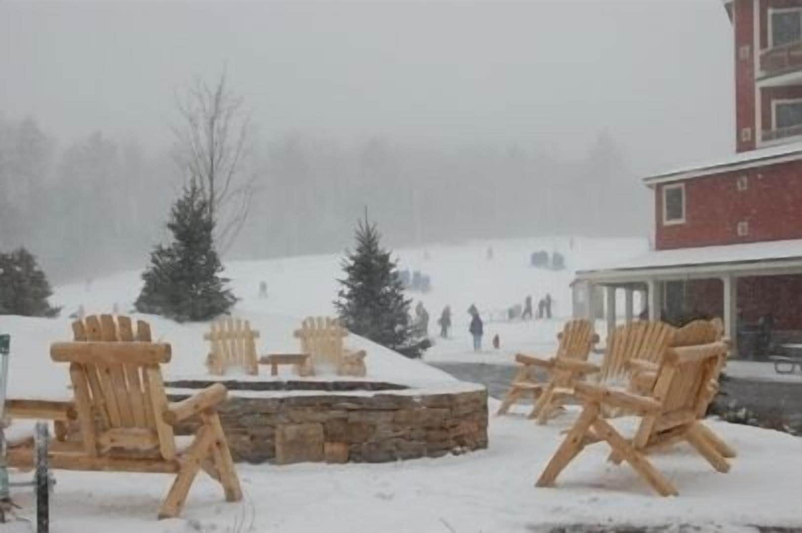 Un espacio exterior cubierto de nieve con sillones Adirondack de madera y un hogar de piedra.