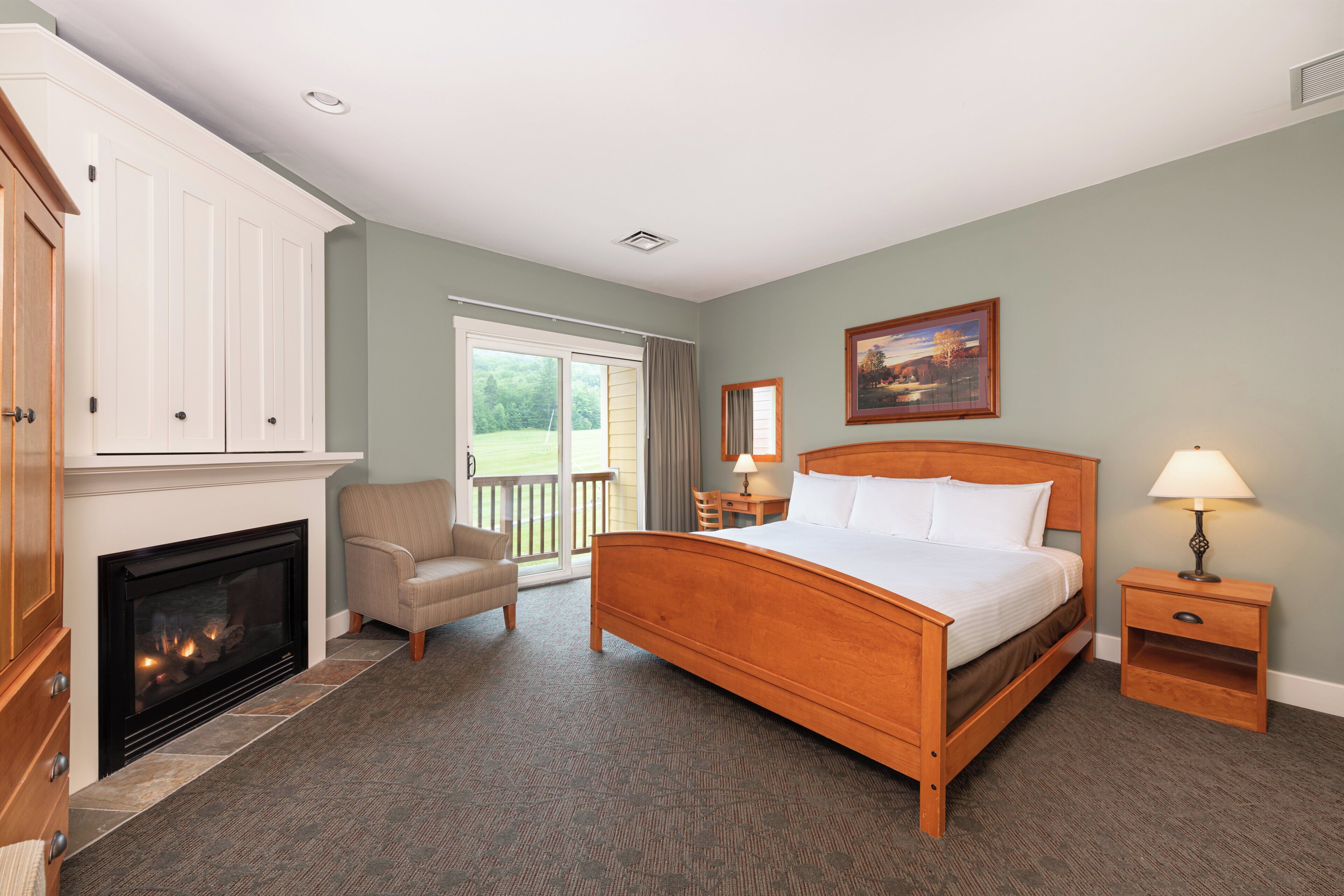 A bedroom with a large bed, a fireplace, a chair, a nightstand, and a view of the outdoors.