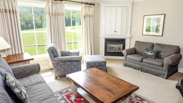 Una sala de estar con sofá, sillón, reposapiés y una mesa de centro de madera. Hay una chimenea y una ventana con cortinas.