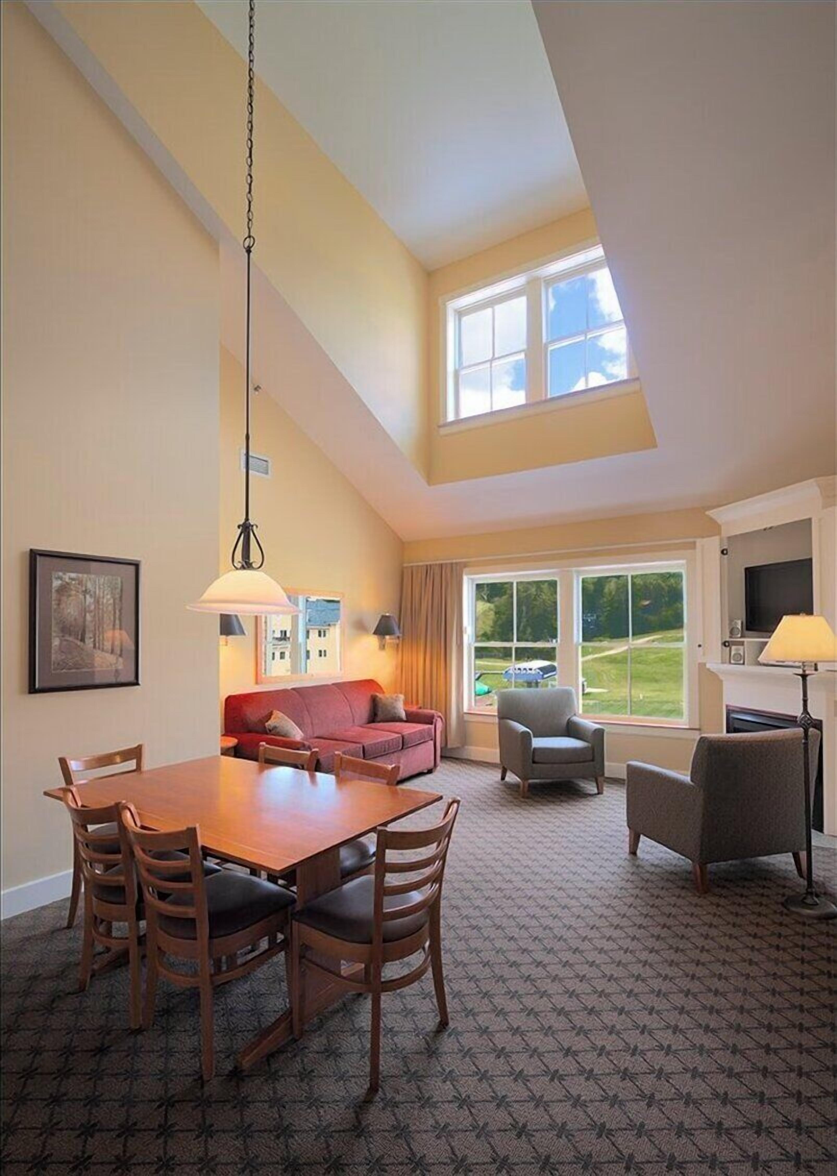 A spacious living room with a high ceiling, a dining area with wooden furniture, and large windows offering a view of the outdoors.
