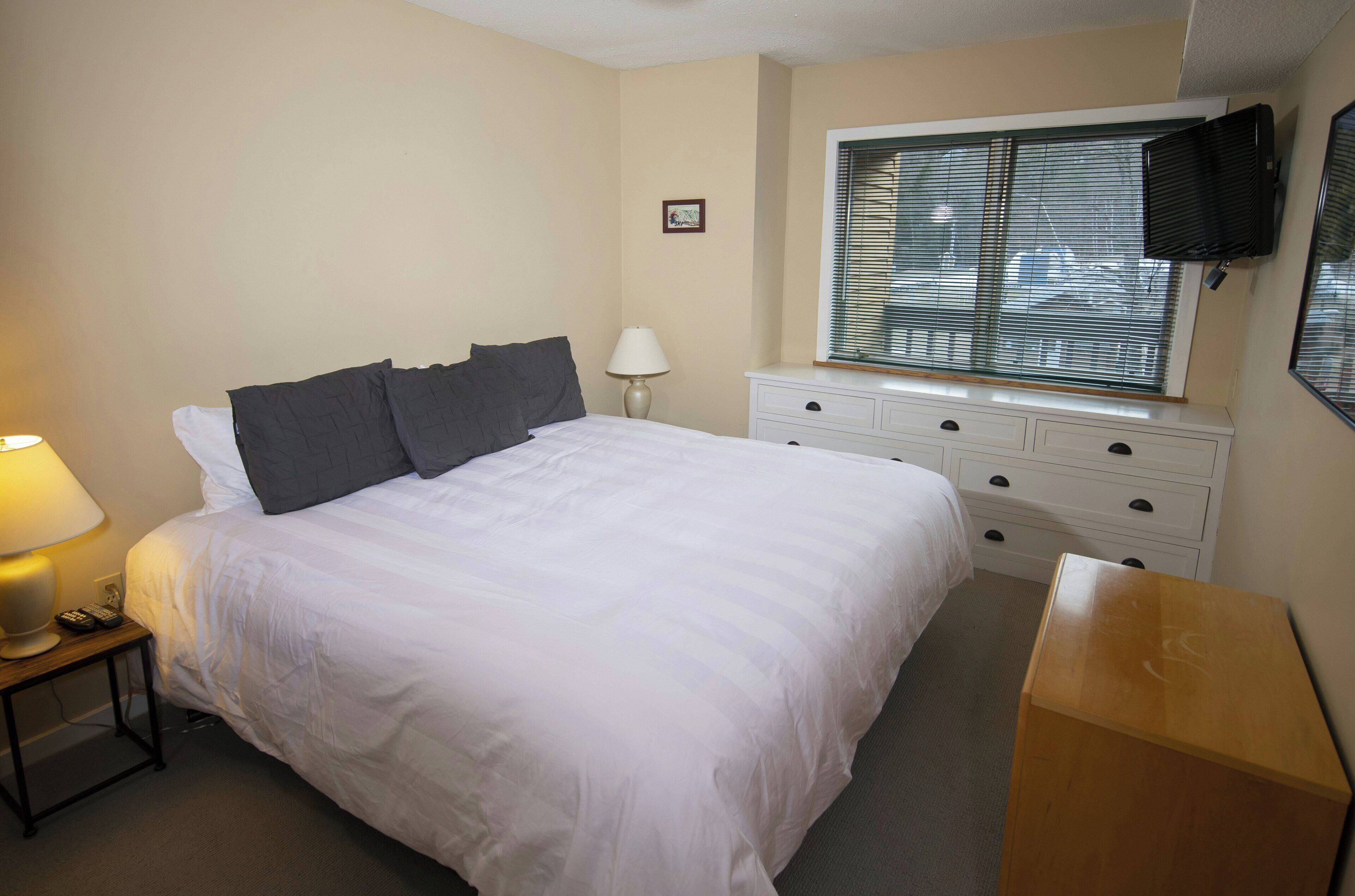 A bedroom with a bed, bedside table, lamp, dresser, window, and TV.