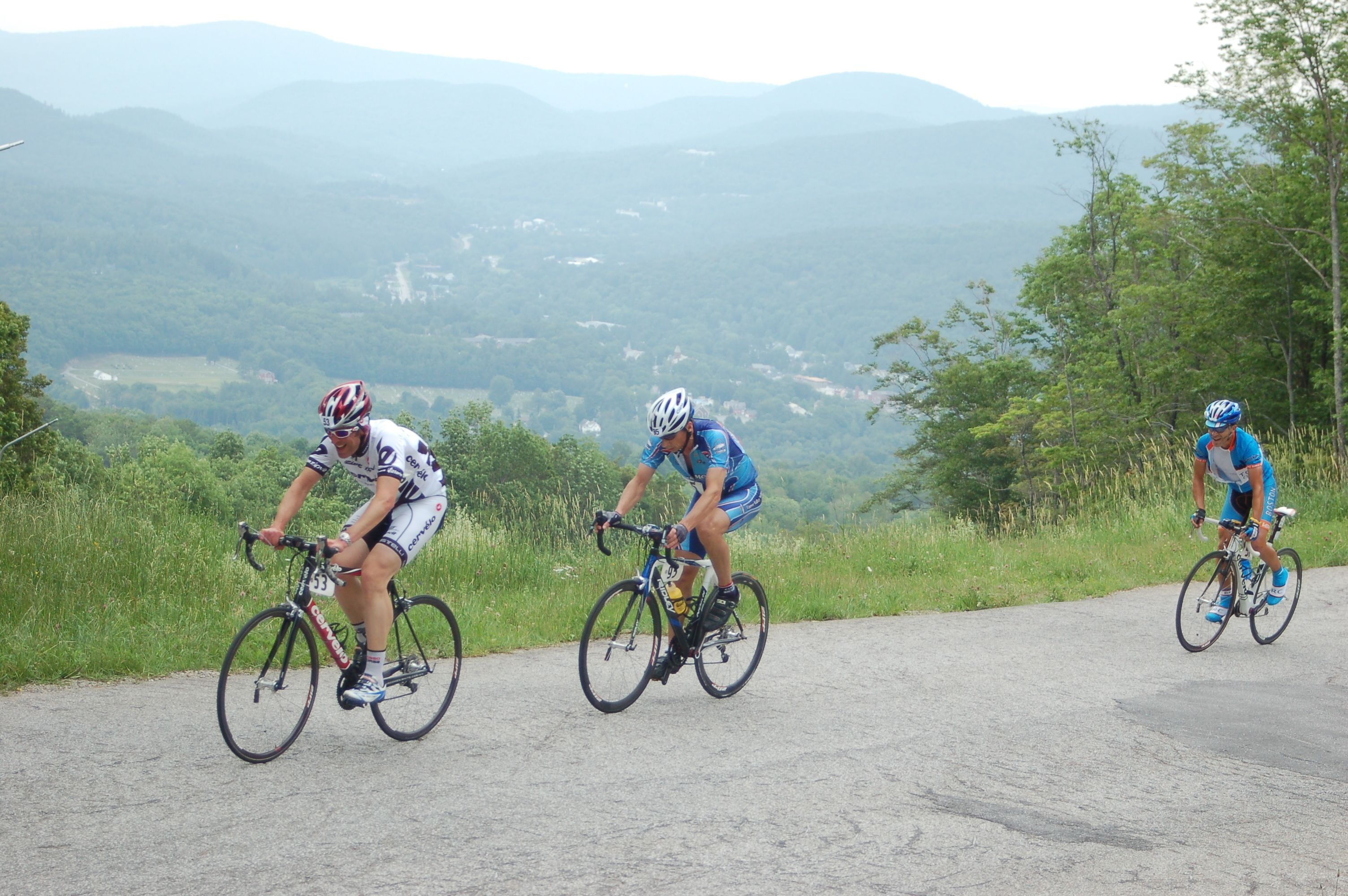 Trois cyclistes sur une route asphaltée, avec une vue panoramique sur les montagnes et la verdure en arrière-plan.