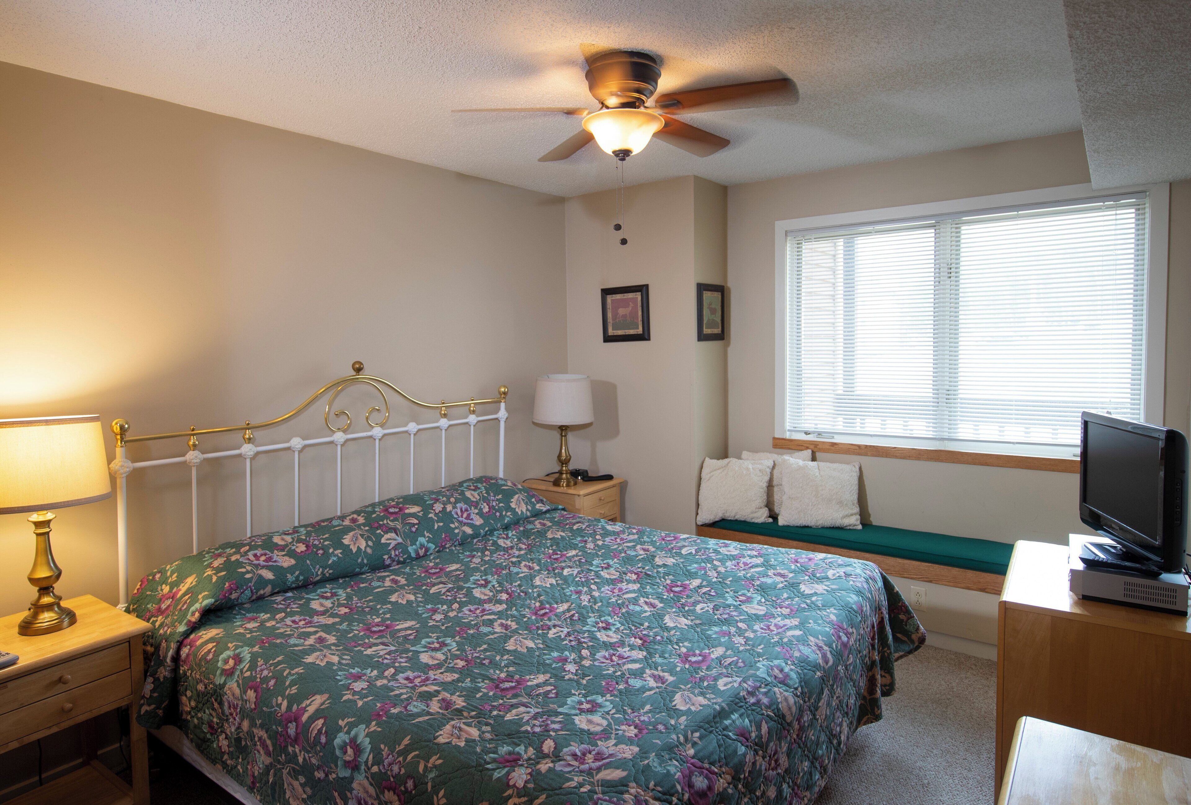 A bedroom with a bed, bedside table, lamp, television, and a window with blinds.