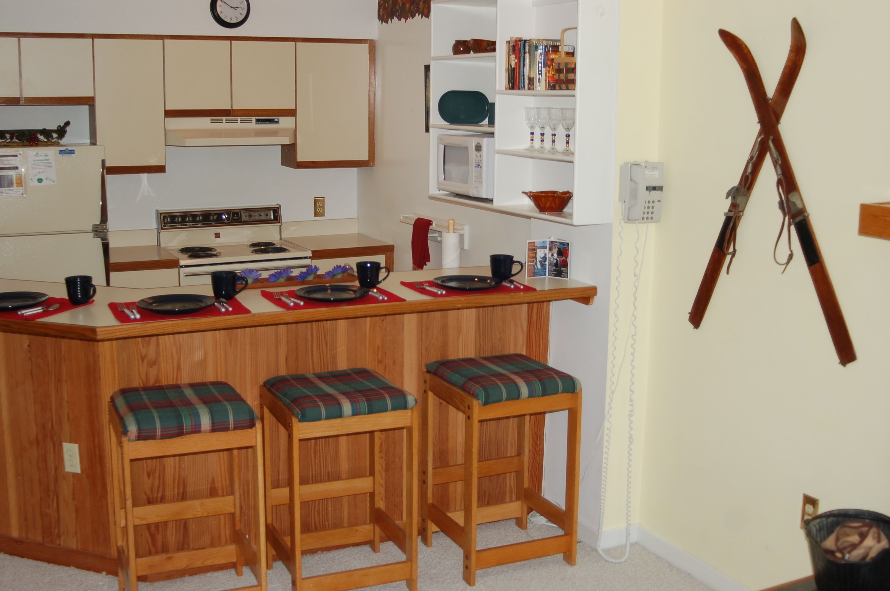 Una cocina compacta con una isla central, taburetes de madera, un microondas y un reloj en la pared.