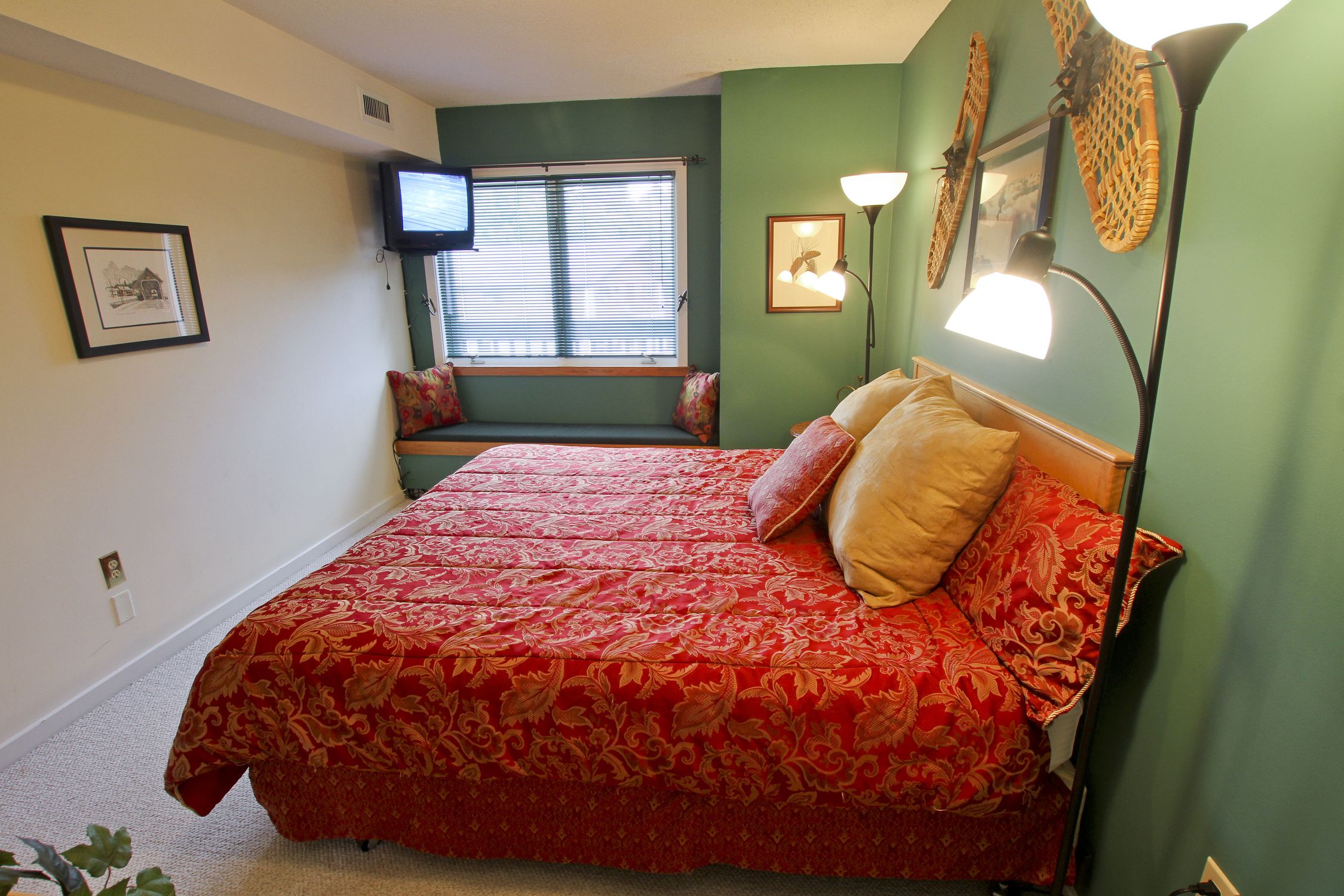 Een slaapkamer met een groot bed, een raam met jaloezieën, een televisie aan de muur en een lamp.