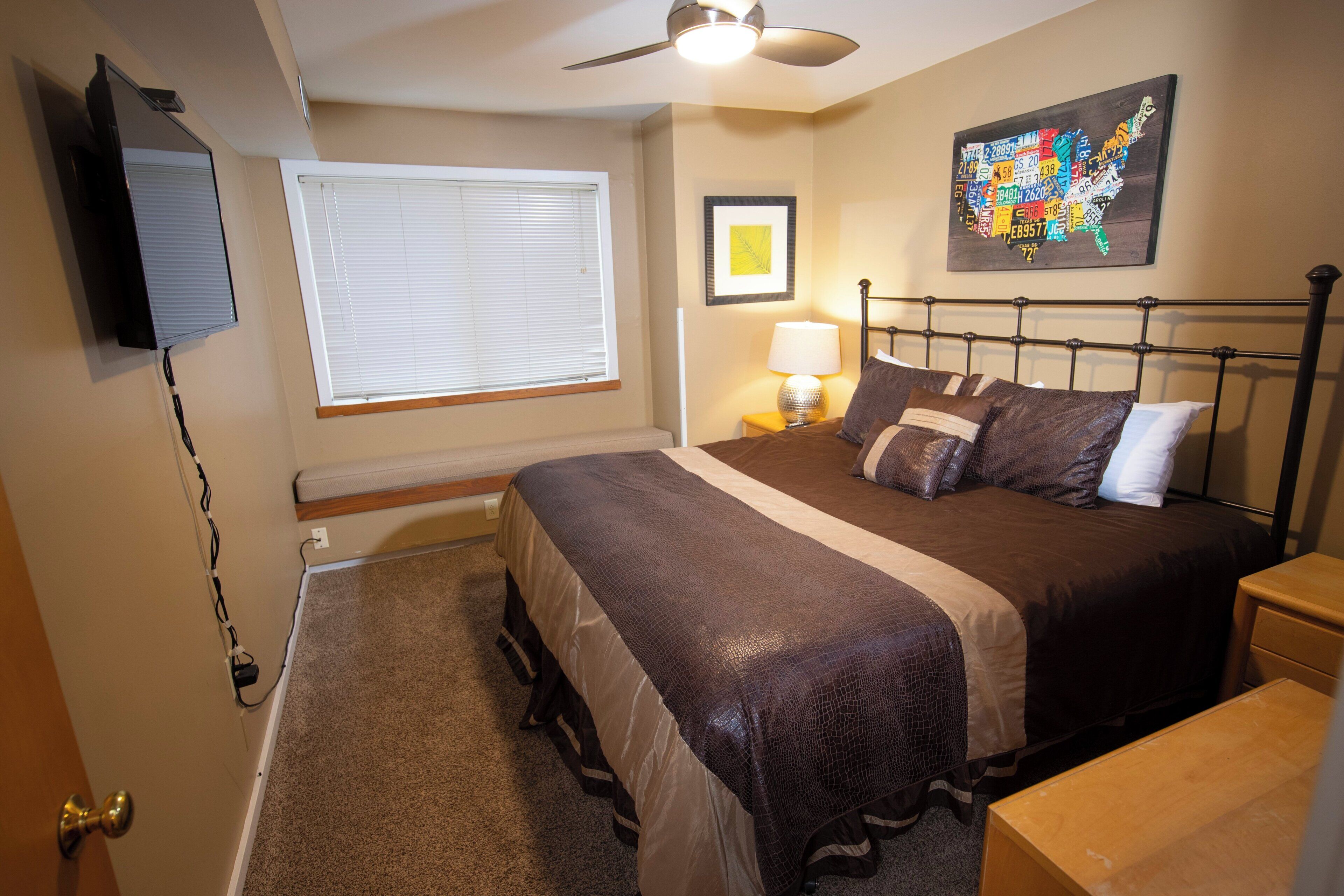 A bedroom with a bed, a ceiling fan, a wall-mounted TV, a window with blinds, a framed picture, and a map on the wall.