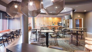 Un moderno vestíbulo de hotel con varias zonas de estar, incluidas mesas y sillas, y una cocina visible al fondo.