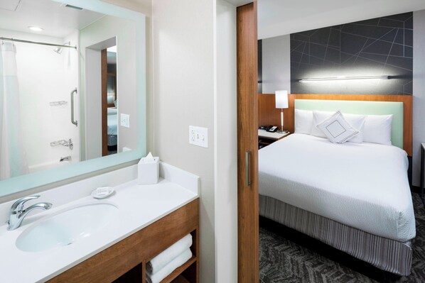 A hotel room with a bathroom featuring a sink, mirror, and shower. The room has a bed with a headboard, pillows, and a bedside table with a lamp.