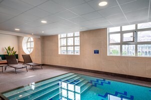 Piscina com azulejos azuis translúcidos, duas espreguiçadeiras e grandes janelas com vista para edifícios.