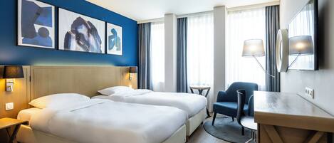 Una habitación de hotel con dos camas, un escritorio, una silla, un televisor y obras de arte en la pared.