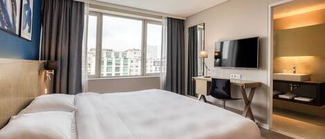 A hotel room with a large bed, a desk with a chair, a flat-screen TV, a bathroom with a sink and mirror, and a city view through the window.