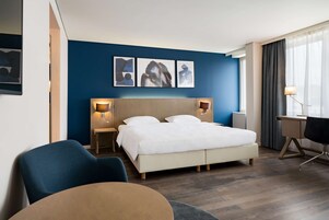 Quarto de hotel com cama, mesas de cabeceira, escrivaninha e uma cadeira.