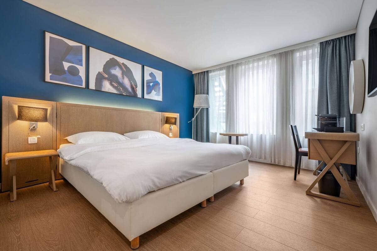 Et hotelværelse med en stor seng, sengeborde, et skrivebord og en stol. Tre billeder hænger på væggen over sengen.