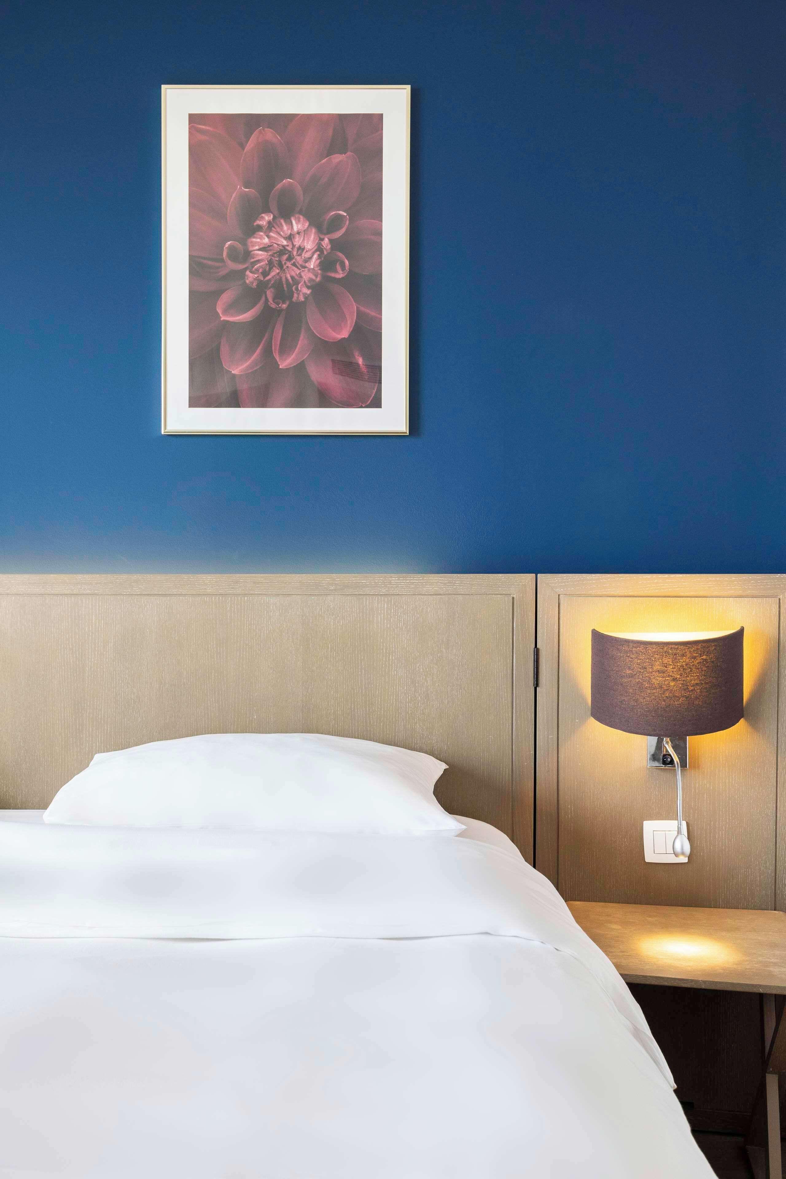 A hotel room with a bed, a wall-mounted lamp, and a framed floral artwork.