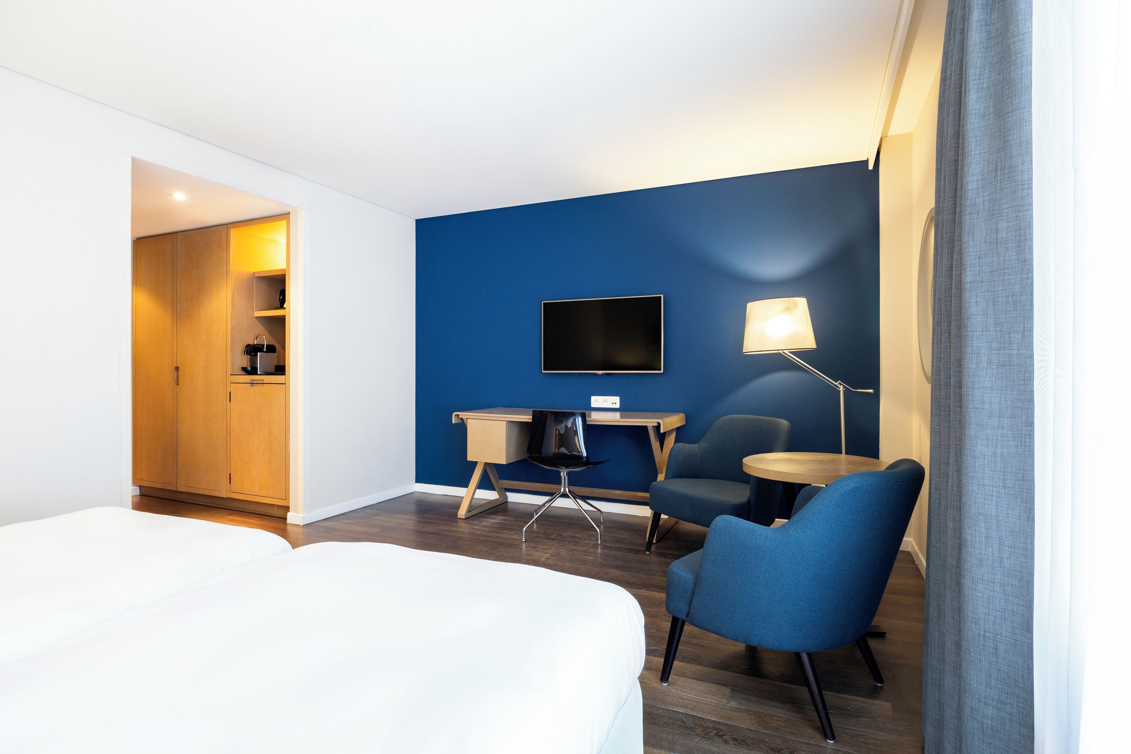 A hotel room with a bed, a desk with a chair, a TV, and a lamp.