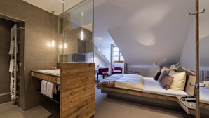 A modern bedroom with a loft-style bed, a wooden bedside table, a glass shower enclosure, and a view of the city through the skylight. - Boutique-Hotel Alemannenhof (Hinterzarten)