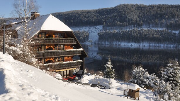 A chalet-style building with a snow-covered roof, multiple balconies, and a view of a lake and forested mountains. - Boutique-Hotel Alemannenhof (Hinterzarten)