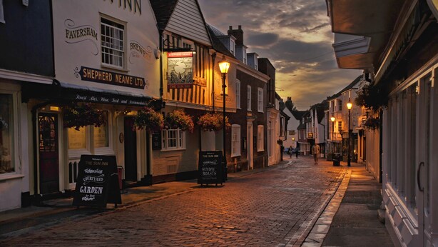 A cobblestone street lined with traditional buildings, including a pub named 'Shepherd Neame Ltd.' with a sign for 'The Sun Inn' and 'Sunday Roasts'. - The Sun Inn (Faversham)