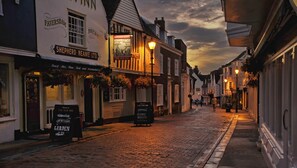 Una calle adoquinada flanqueada por edificios tradicionales, incluyendo un pub llamado 'Shepherd Neame Ltd.' con un letrero que indica 'The Sun Inn' y 'Asados Dominicales'.