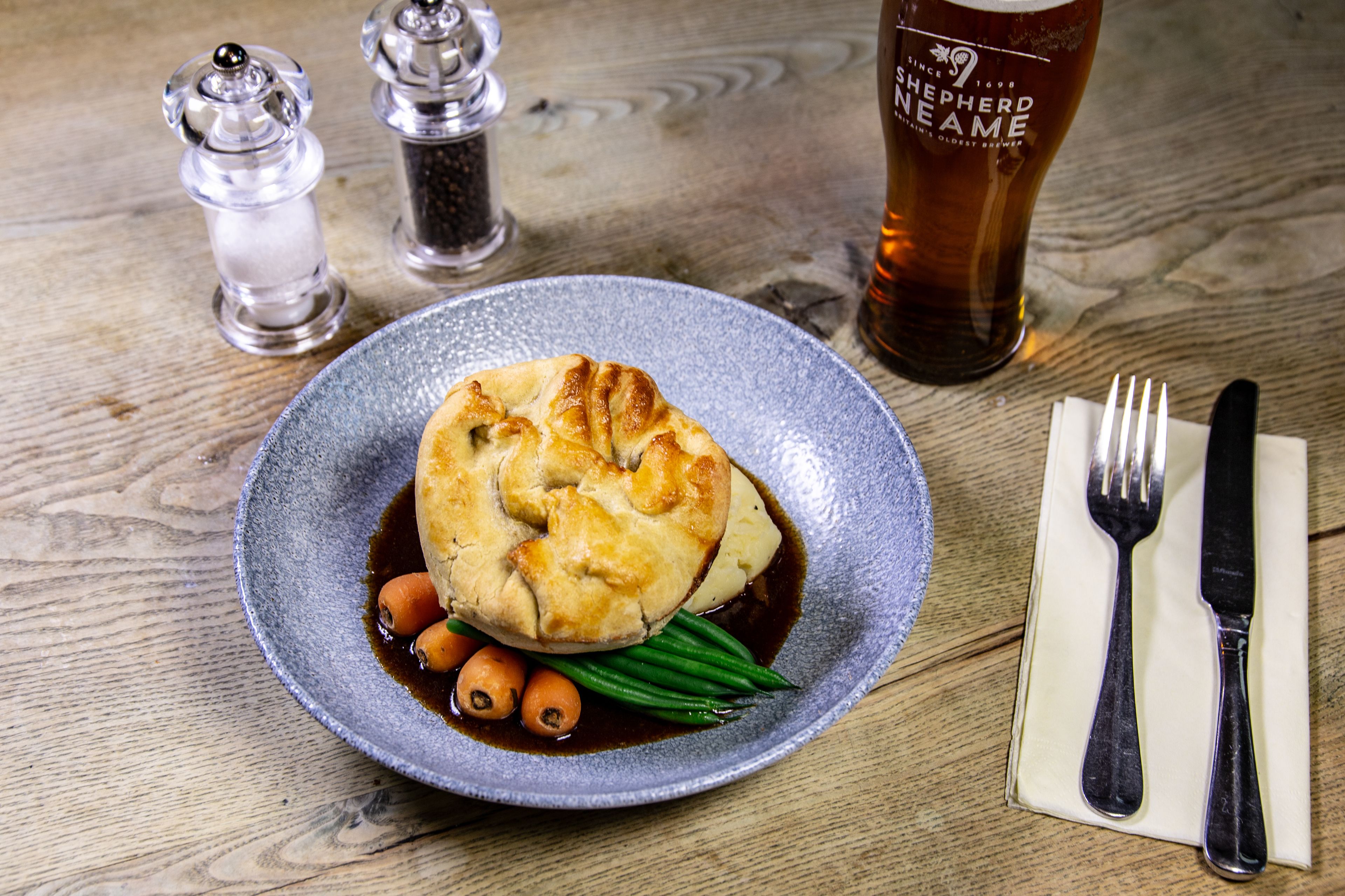 A plate of food with a pastry, green beans, and carrots, accompanied by a glass of beer and salt and pepper shakers on a wooden table.