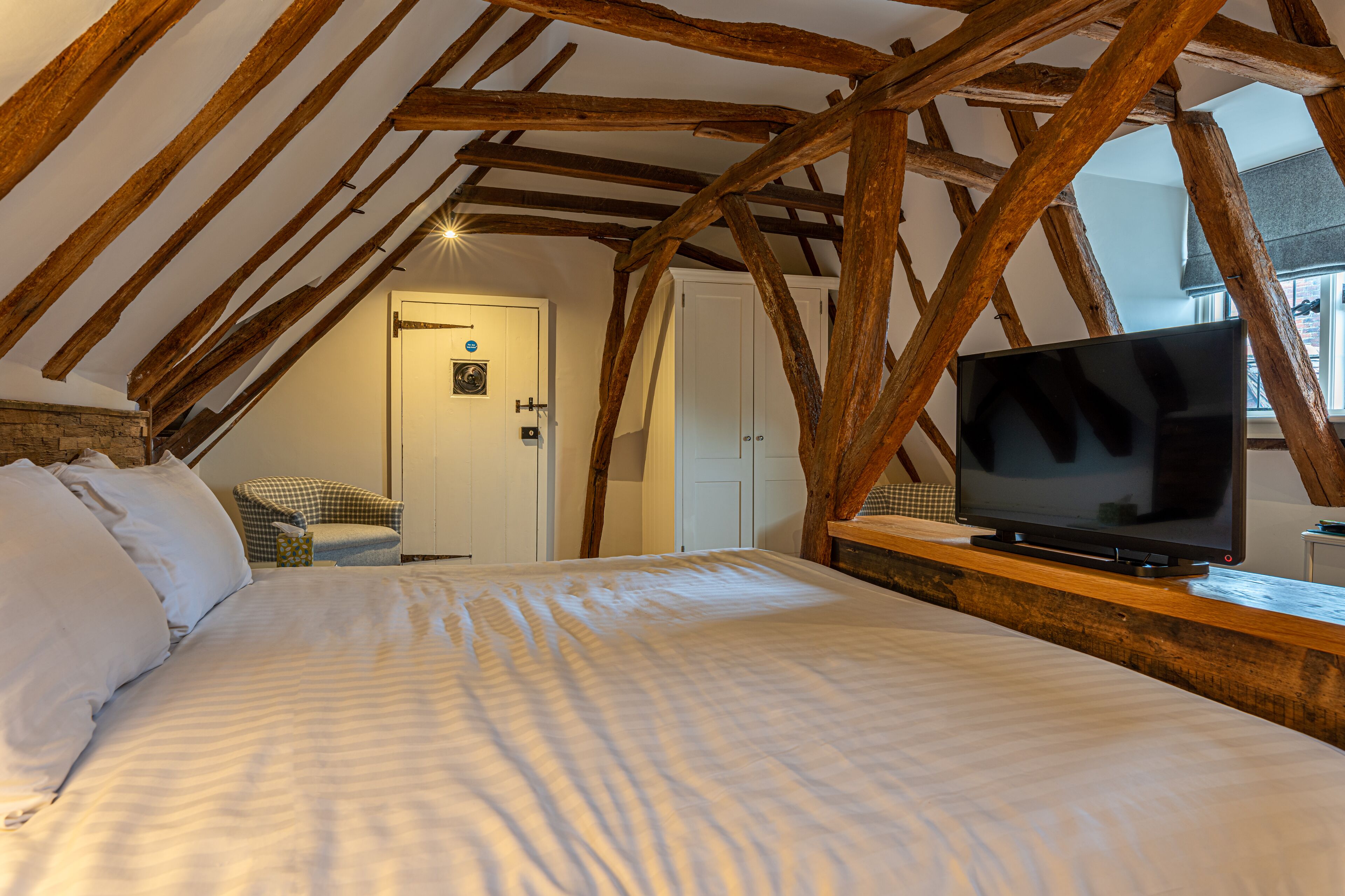 Quarto com uma cama grande, vigas de madeira, uma televisão sobre uma cômoda de madeira e uma porta ao fundo.