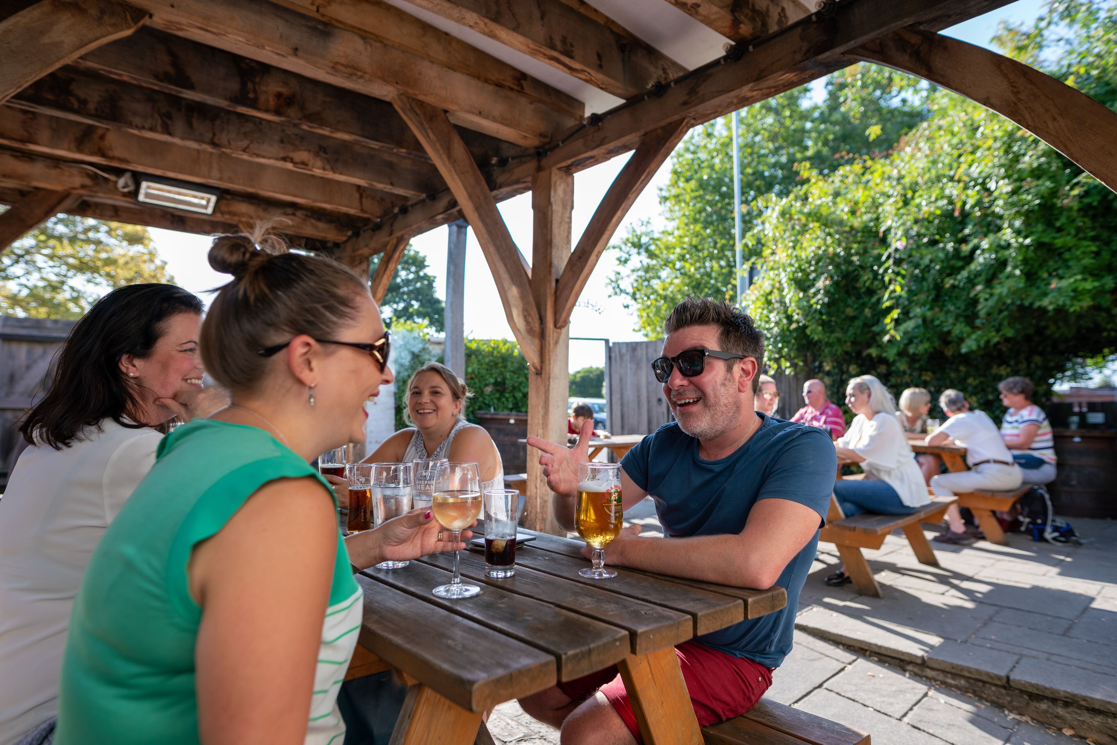 Un grupo de personas sentadas en una mesa de madera al aire libre, con bebidas, bajo un pabellón de madera.