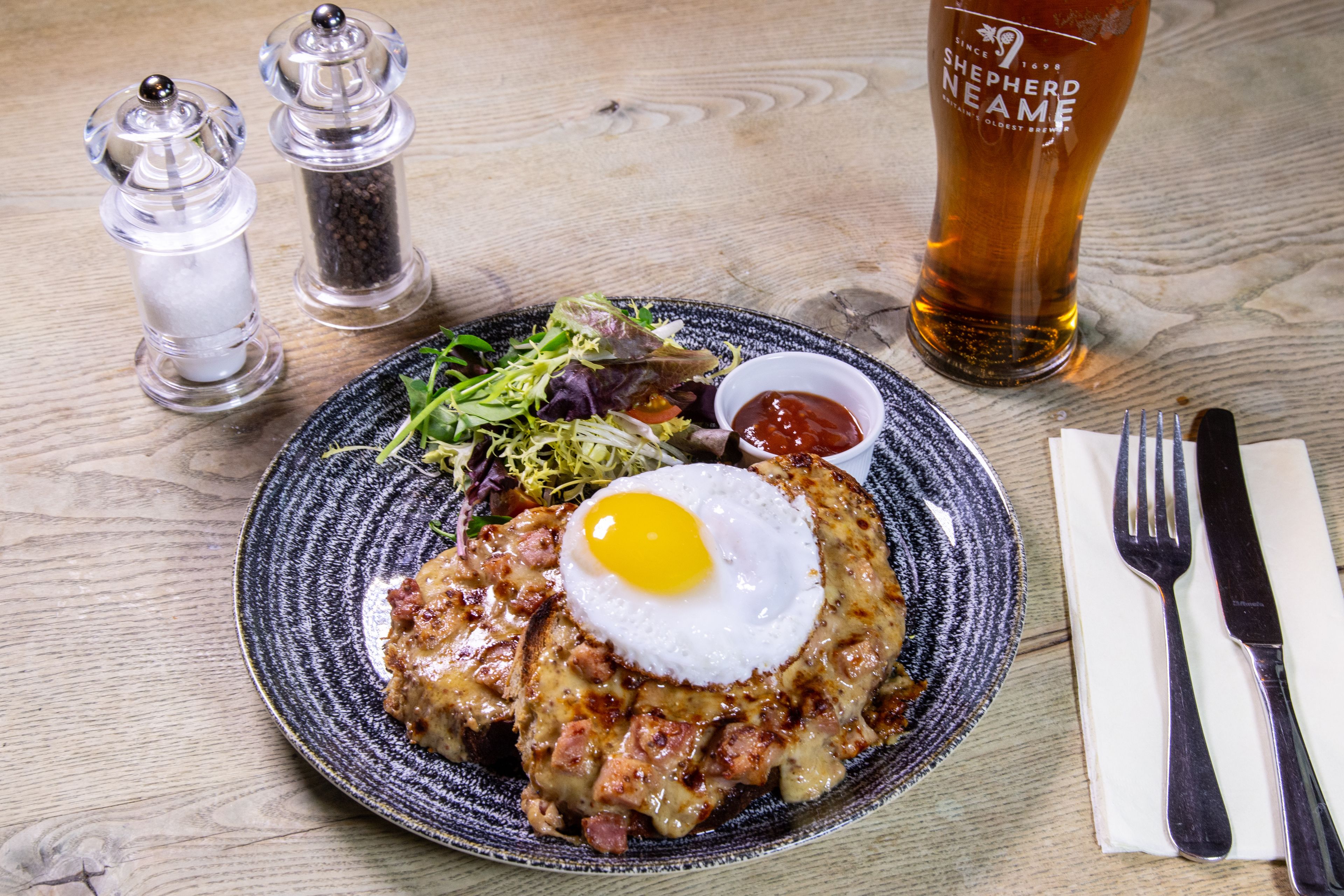 Een bord met een gebakken ei, hash browns en een salade, vergezeld van een glas bier en zout- en peperstrooiers op een houten tafel.