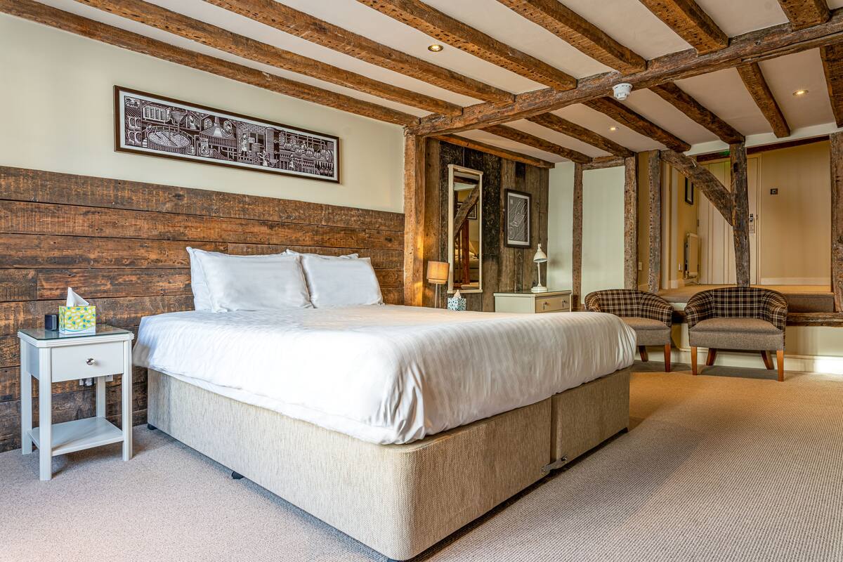 A bedroom with a large bed, wooden beams, a nightstand, a chair, and a mirror.