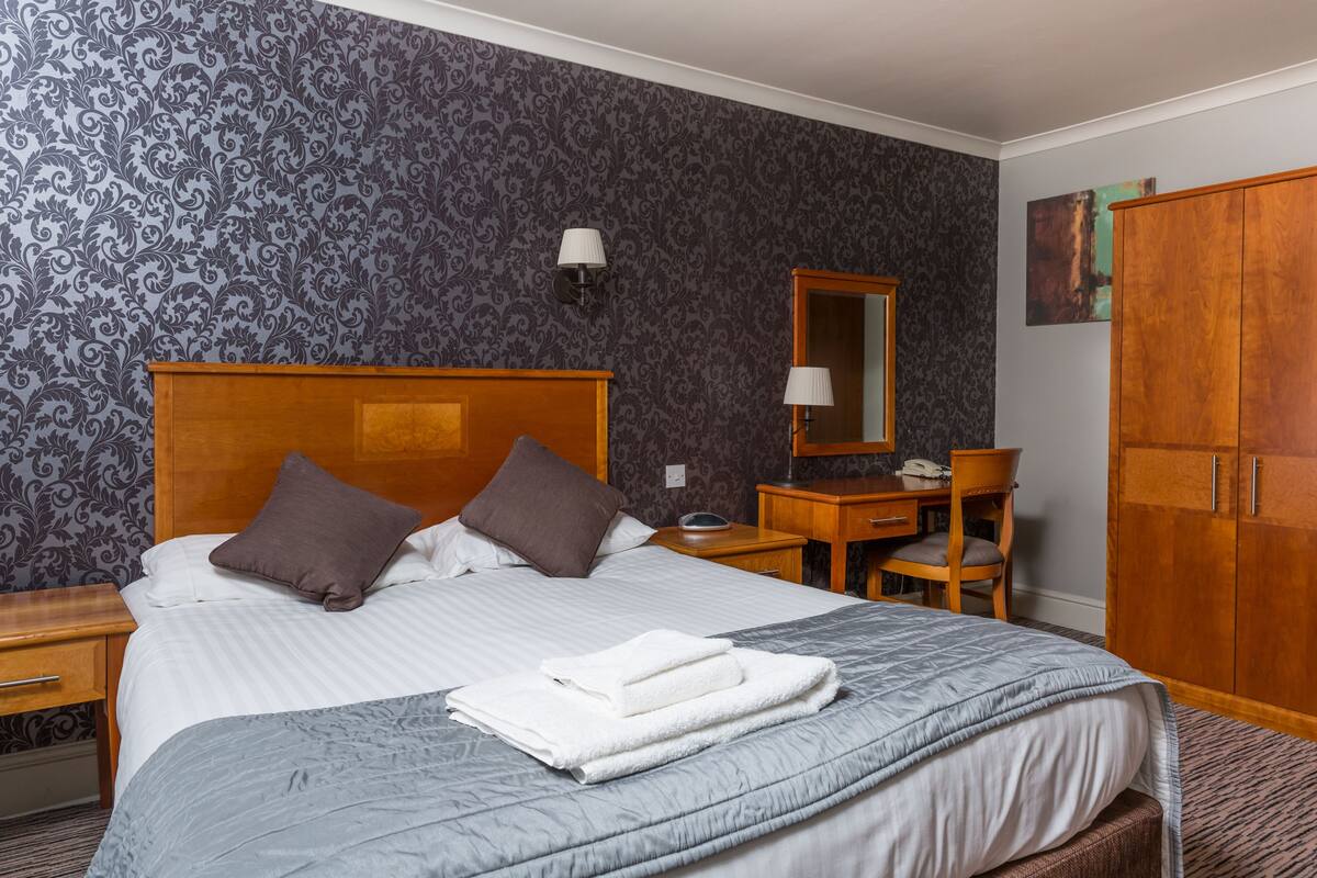 A hotel room with a large bed, a wooden headboard, a desk with a chair, a wardrobe, and a mirror.