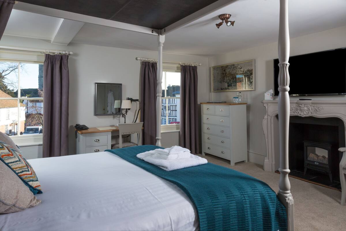A four-poster bed with a teal throw blanket, a white dresser, a mirror, a television, and a fireplace.