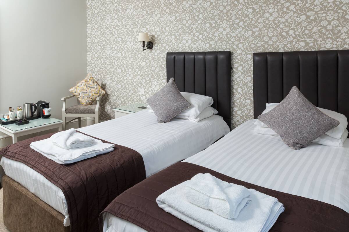 A hotel room with two beds, each with a brown headboard, white linens, and a grey pillow. There is a small table with a kettle, cups, and a tray of towels.