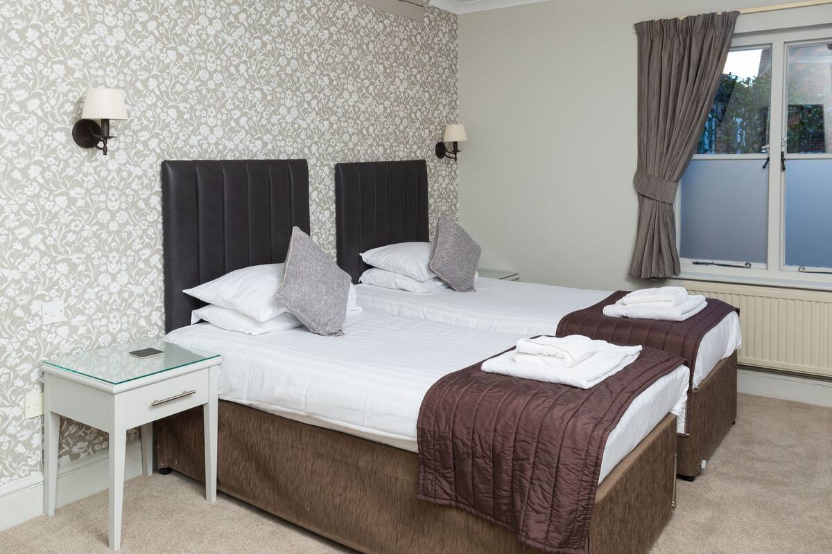 A double bed with a white duvet and two pillows, a bedside table with a lamp, and a window with curtains.