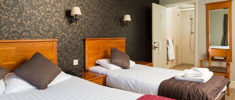 Habitación de hotel con dos camas, cabecera de madera, mesita de noche, lámpara, espejo y puerta de baño.