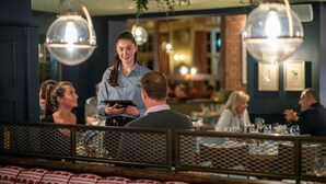 A restaurant interior with a waiter holding a tablet, tables with glasses and cutlery, and hanging spherical light fixtures. - The Crown Inn (Chislehurst)