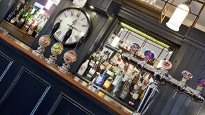 A bar area with a large clock, multiple beer taps, and various bottles of alcohol. - The Millers Arms (Canterbury)