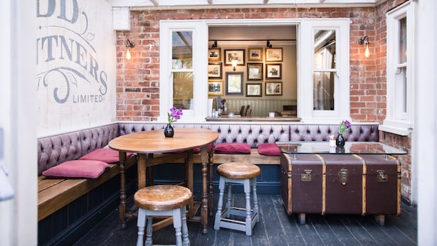 A cozy corner with a round wooden table, cushioned bench seating, and a vintage suitcase. - The Millers Arms (Canterbury)