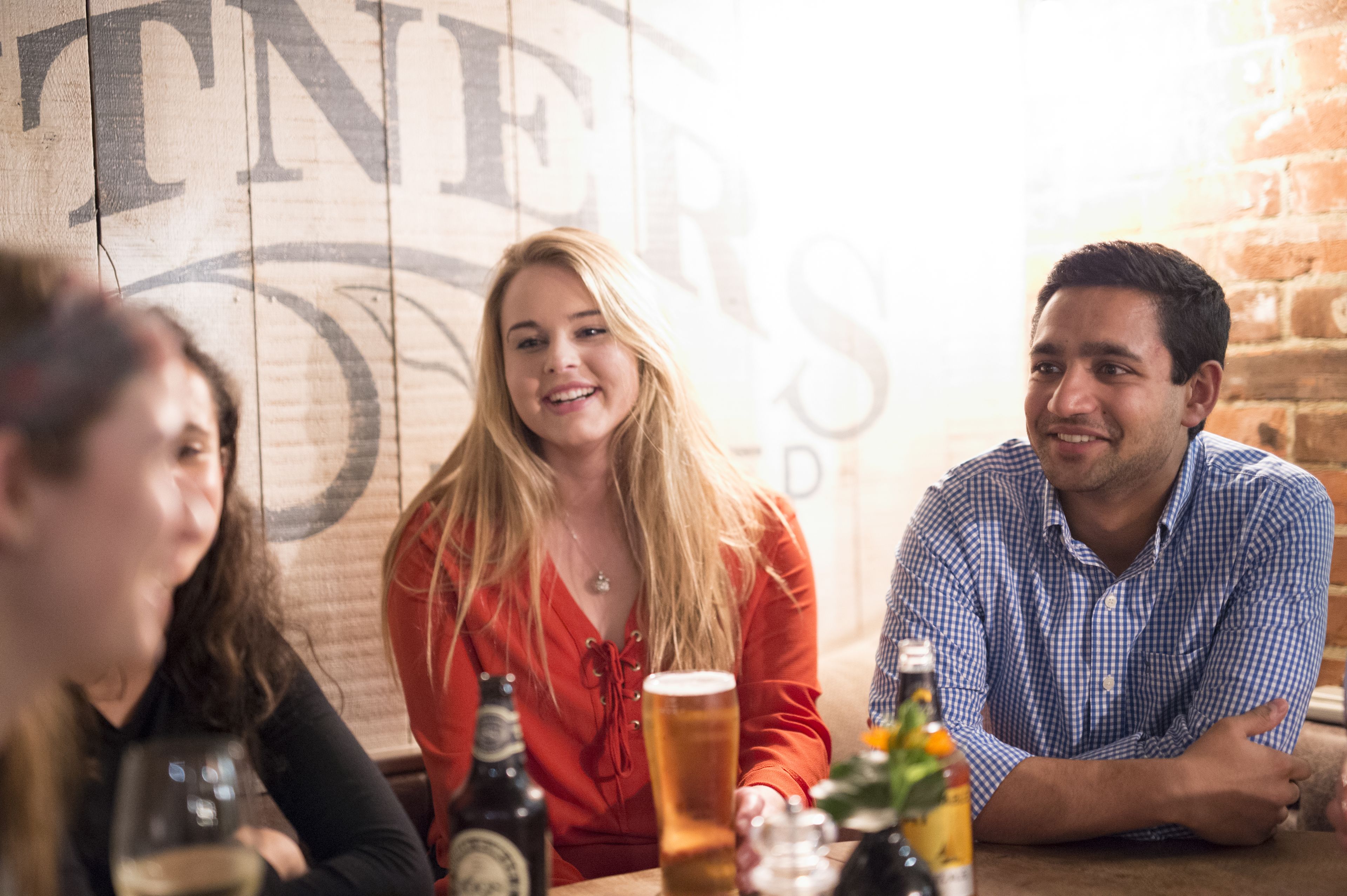 Eine Gruppe von Personen sitzt an einem Tisch mit Bierflaschen und Gläsern in einem Raum mit Backsteinwand und einem großen Schild mit teilweise lesbarem Text.