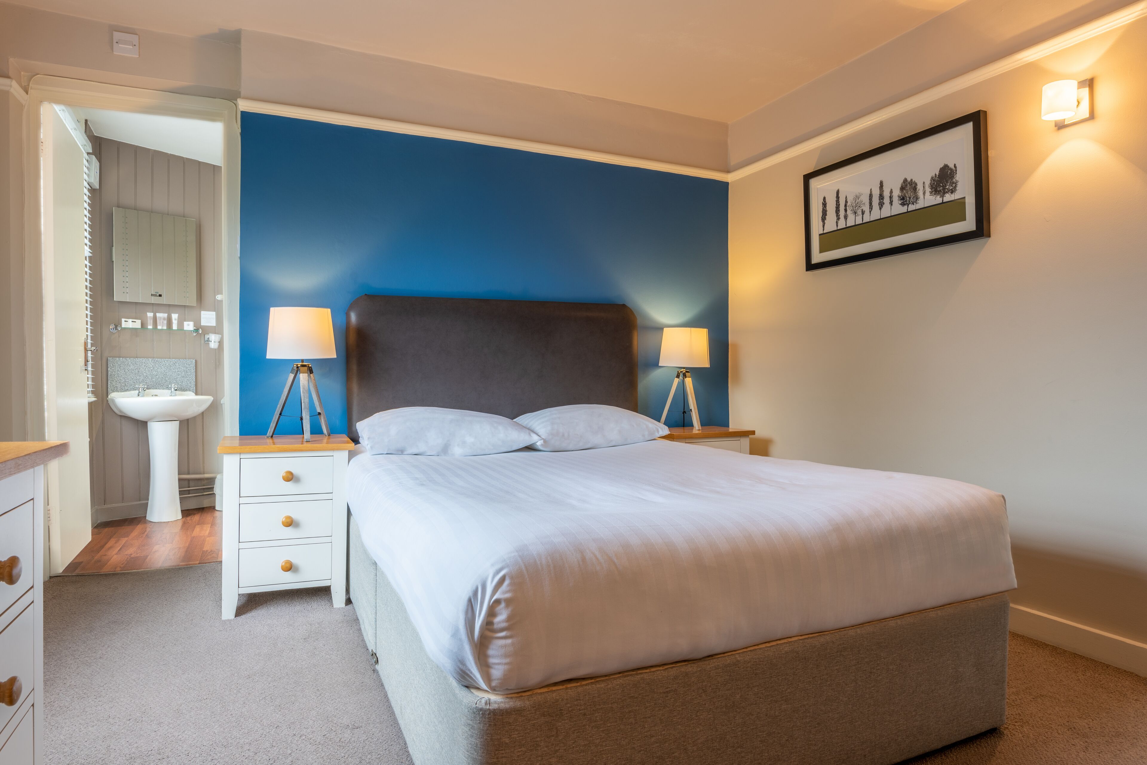 A bedroom with a large bed, bedside tables, a nightstand, a wall-mounted lamp, and a framed picture.