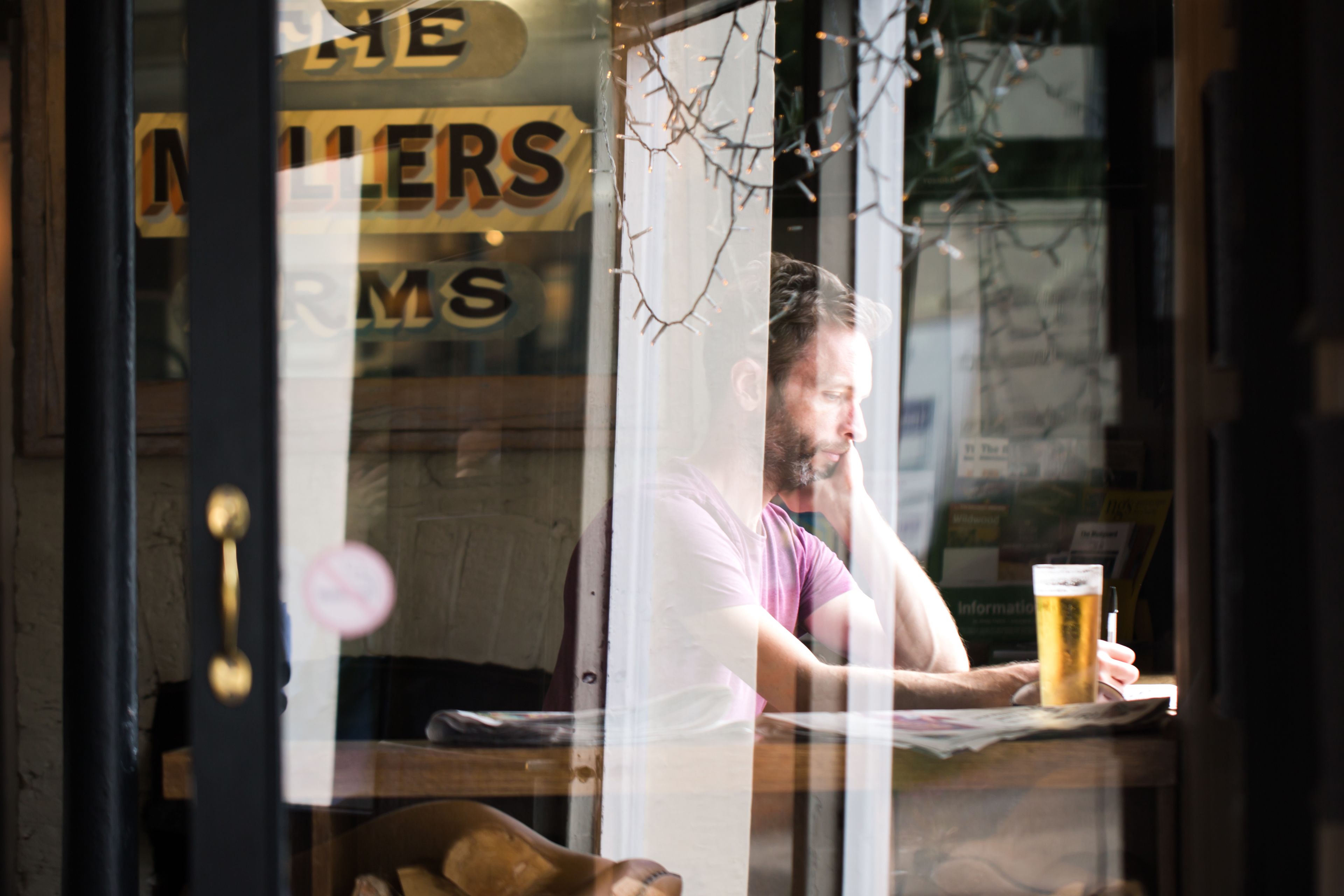 A person sitting at a table with a beer in front of them, inside a building with a sign that reads "THE MALLERS ROOM".
