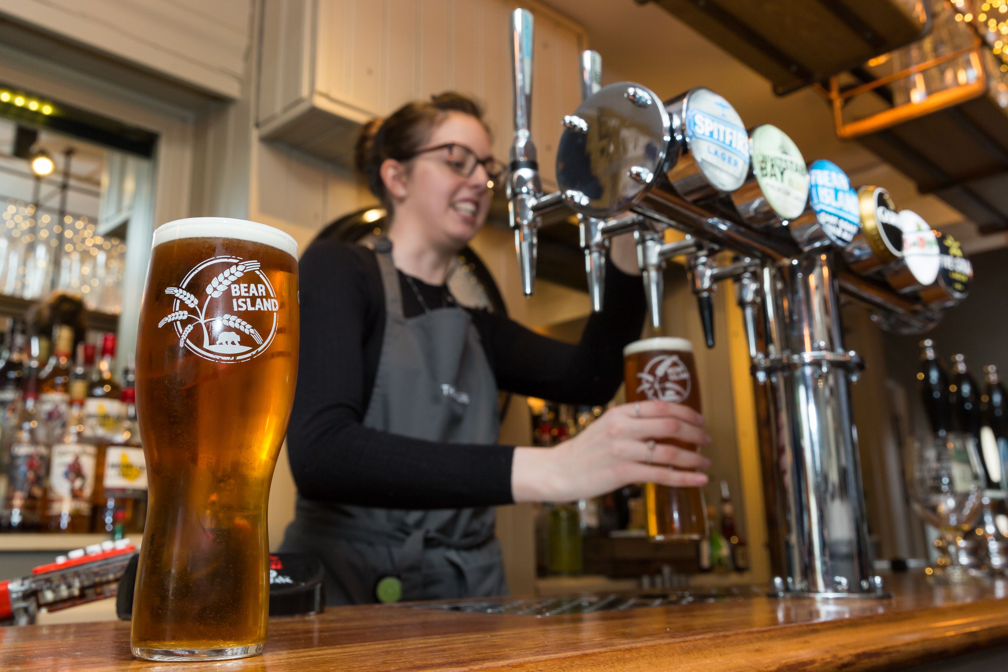 Un barman se tient derrière le bar, où l’on peut voir une tireuse à bière avec l’inscription « Bear Island » et un grand verre à bière.